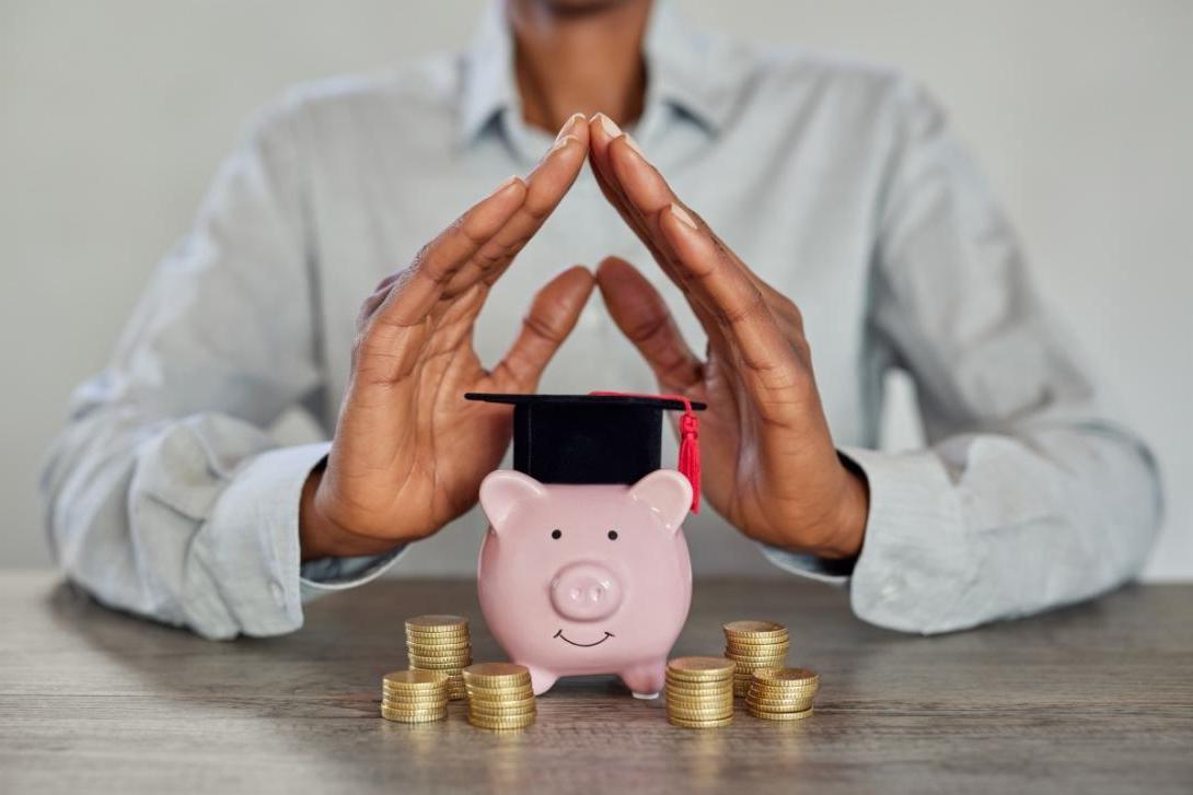 Hands held over a small piggy bank wearing a commencement cap with money stacks around it